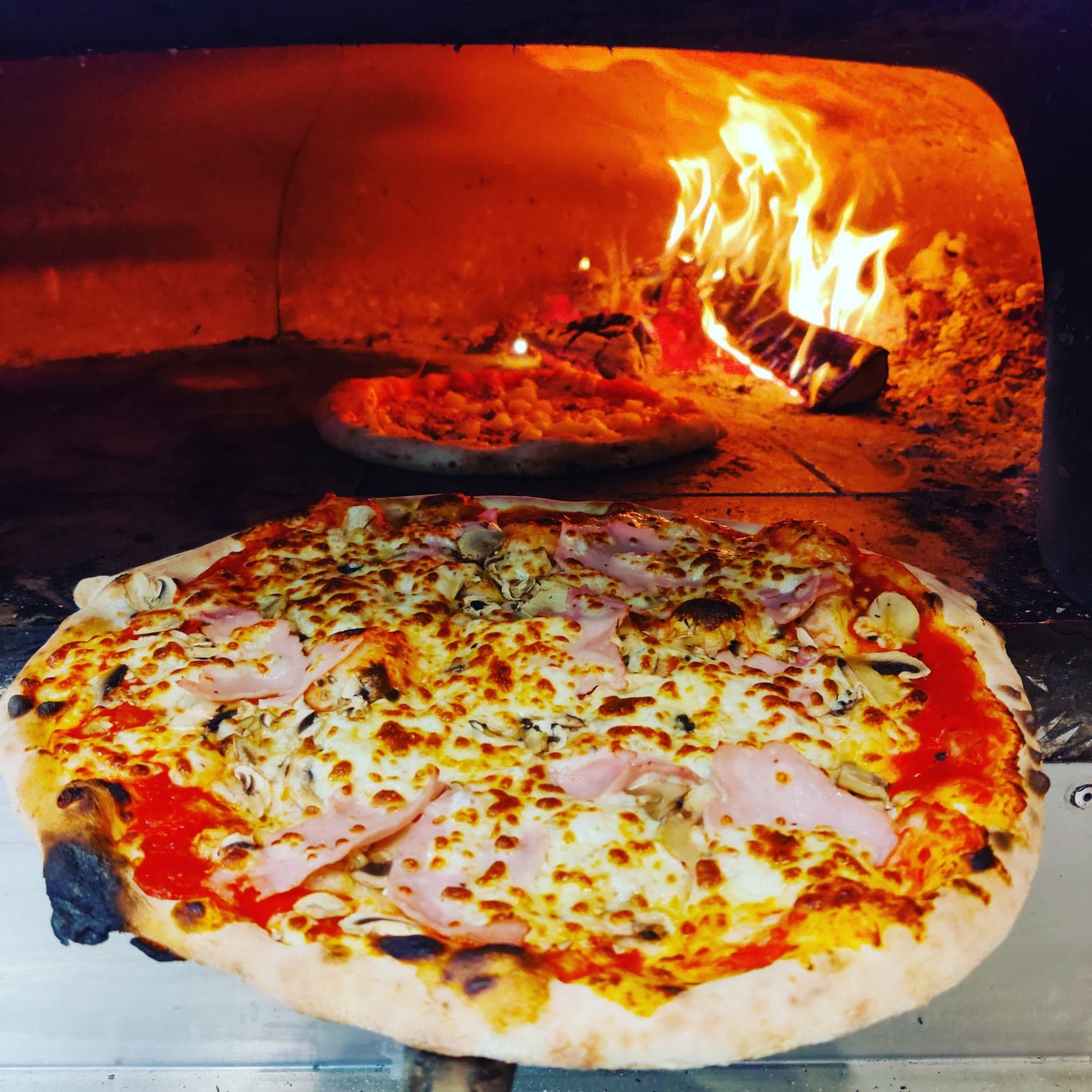 Une pizza au jambon et aux champignons sur la pelle, devant les flammes du four.