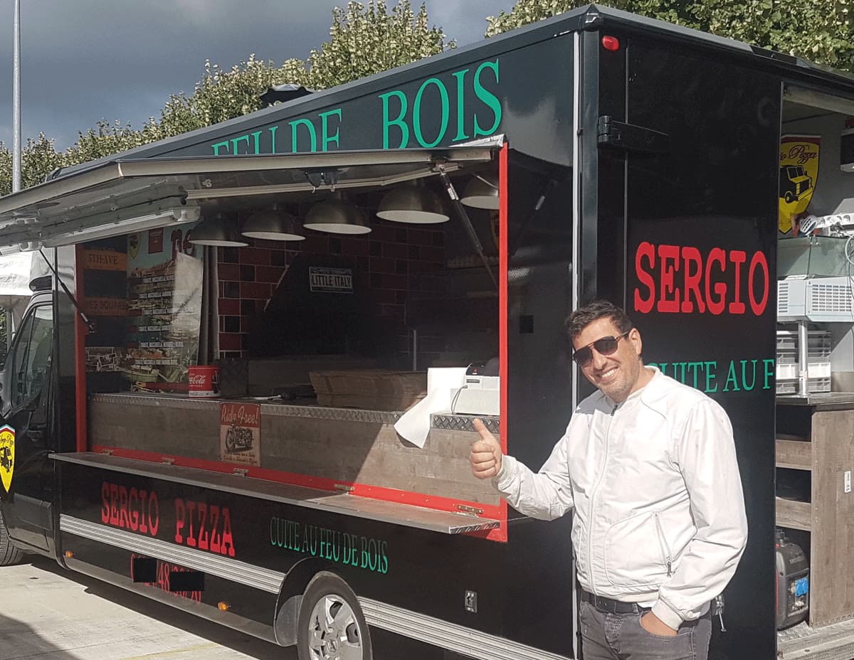 Sergio, pouce levé, devant le camion noir aux lettres rouges et vertes.